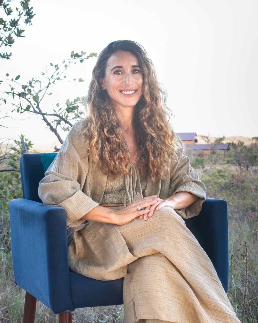 Me in a deep blue armchair outdoors, in linen, hands folded, smiling into the light.