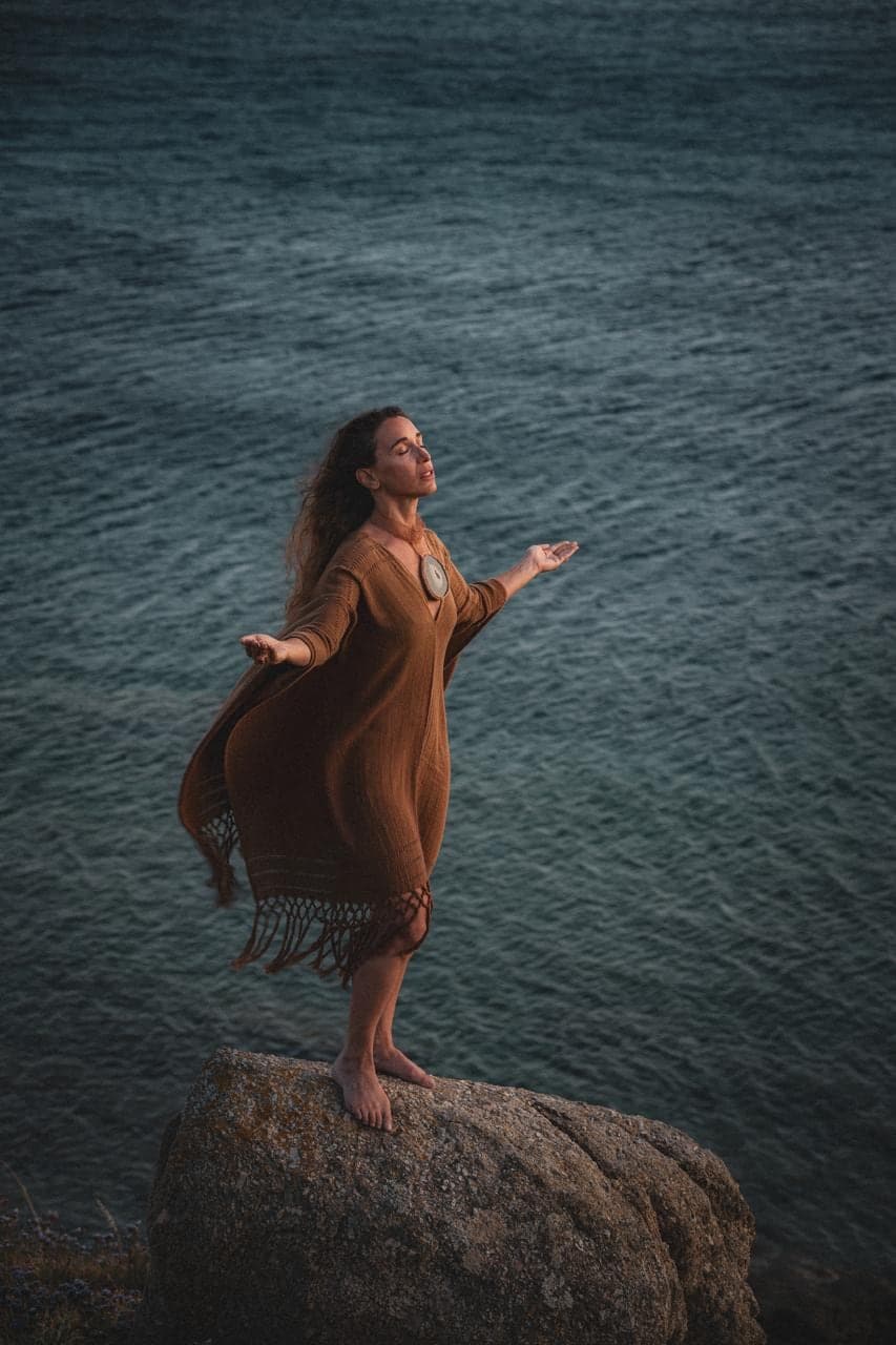 Standing on a rock over dark water, arms open, the ochre wrap moving in the wind.